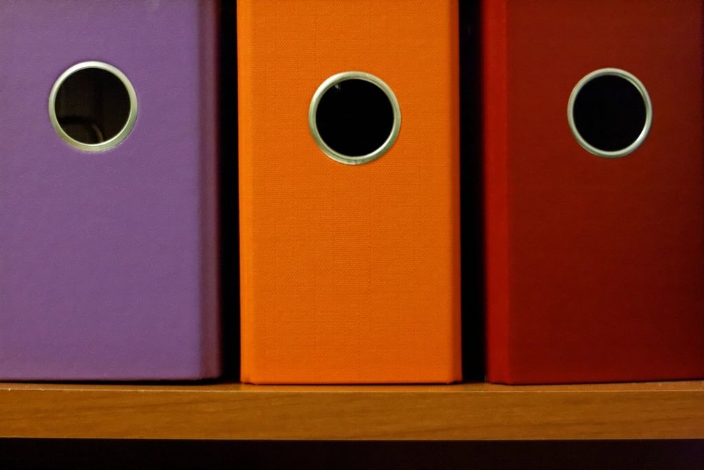 Trois classeurs de couleur, dont un orange, rangés côte à côte sur une étagère en bois