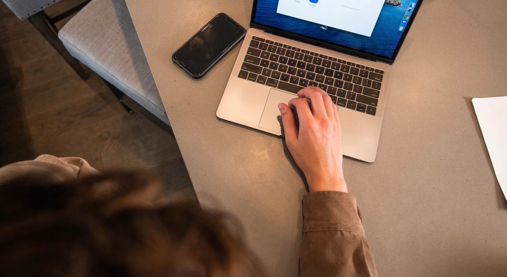 Une personne assise à une table travaille sur un ordinateur portable, son téléphone posé à côté
