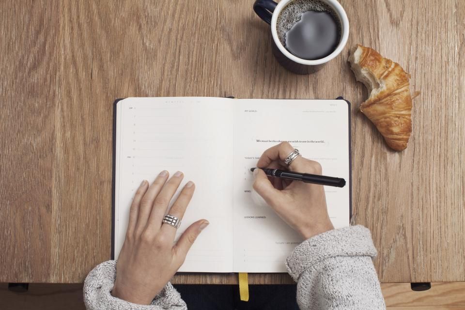 Deux mains qui notent des chiffres dans un carnet ouvert, posé sur une table en bois à côté d'une tasse de café