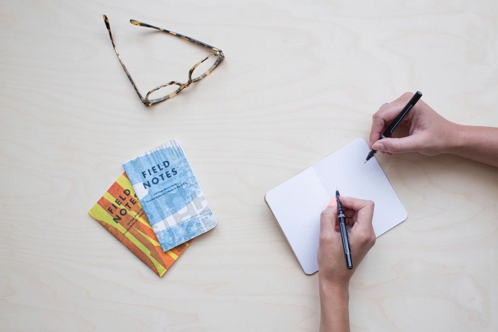 Deux mains prennent des notes dans un carnet posé sur une table claire, à côté de lunettes